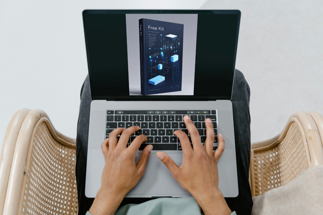 Top-down view of a person typing on a silver laptop while wearing black pants, seated in a woven beige chair. The laptop screen displays a digital cover for a 'Free Kit'.